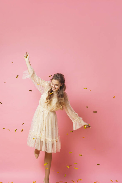 Merry Christmas. A beautiful attractive girl with wavy hair in a light shiny dress dances under golden falling, flying confetti on a pink background in the studio. In full growth. Copy space - Фото, зображення