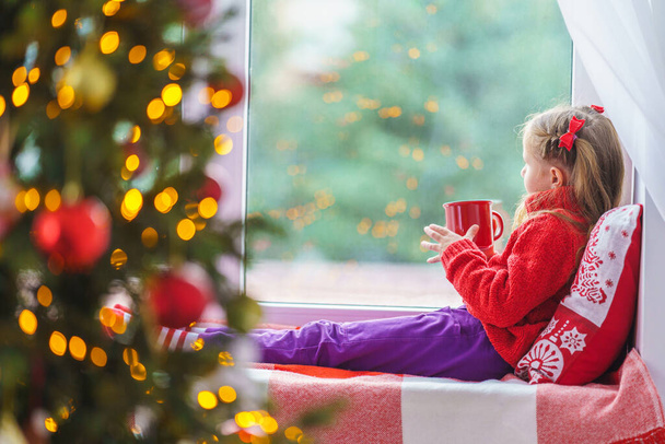 A cute little girl is sitting by the window with a cup of hot drink. Decorated Christmas tree. a child in warm clothes with a blanket and a pillow by the window. Merry Christmas and happy holidays. - Foto, afbeelding