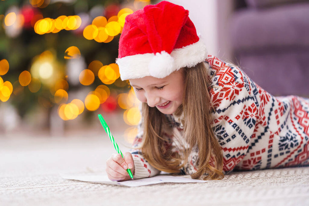a small child with a Santa hat and New Year's pajamas, lying on the floor, writes a letter with wishes to Santa Claus. Christmas order. removed from the floor level. Merry Christmas and happy holidays - Фото, изображение