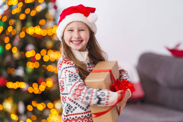 cute cheerful little girl in pajamas and a Christmas hat with a box tied with a red ribbon. he laughs and holds a gift in his hands. Christmas tree in the light of garlands in the background - Foto, Imagem