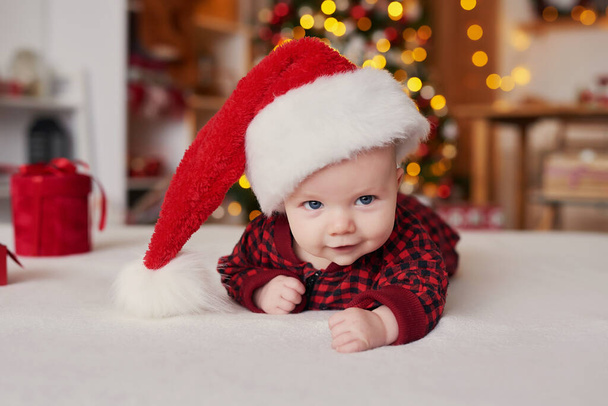 Kerst baby jongen in santa hoed op achtergrond van kerstboom met geschenken - Foto, afbeelding
