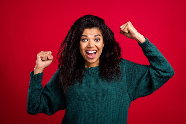 Photo of confident lucky dark skin woman dressed green pullover smiling rising fists isolated red color background - Фото, зображення