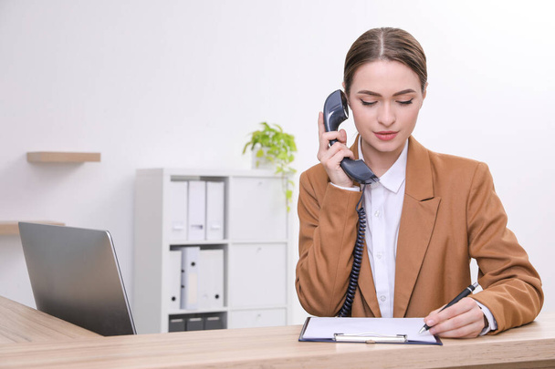 Female receptionist with clipboard talking on phone at workplace. Space for text - Фото, зображення