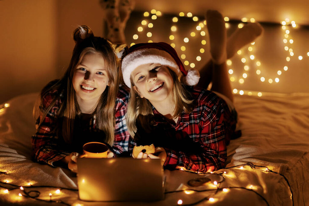 Two cute sisters use a laptop at home by the Christmas tree on Christmas Eve, watch a movie or send Christmas greetings. Winter evening at home with family and children - Fotó, kép