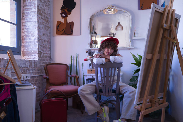 Jonge vrouw, schilder, ontwerper en kunstenaar met een rode baret, zittend op een stoel ondersteboven en leunend op de rugleuning, borstels in de hand in haar atelier. Concept schilderen, tekenen, kunstenaar, ontwerper. - Foto, afbeelding