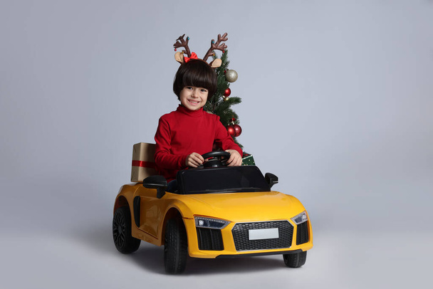 Cute little boy with Christmas tree and gift box driving children's electric toy car on white background - 写真・画像
