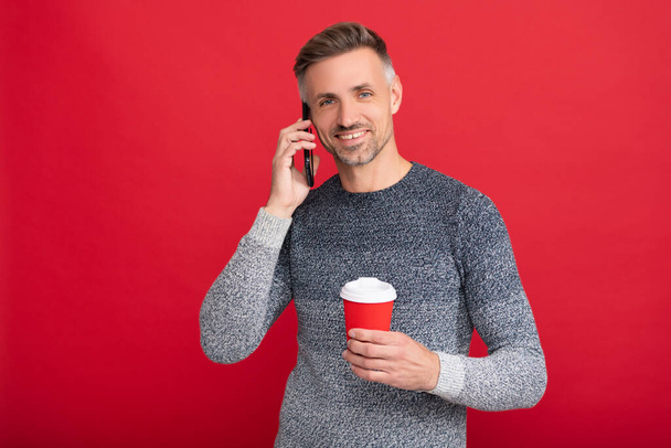 cheerful grizzled guy in sweater drink coffee with smartphone on red background, communication - Photo, Image
