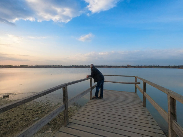 Ein älterer Mann beobachtet an einem Wintertag den Sonnenuntergang auf der Seebrücke. Schöne Landschaft des Ibars Vilasana Sees in Katalonien, Nordspanien. Sonnenuntergang auf dem See - Foto, Bild