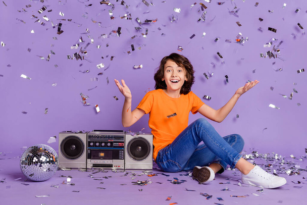 Pleine longueur taille du corps photo heureux garçon assis près de magnétophone disco ball avec confettis volants isolé fond de couleur pourpre - Photo, image