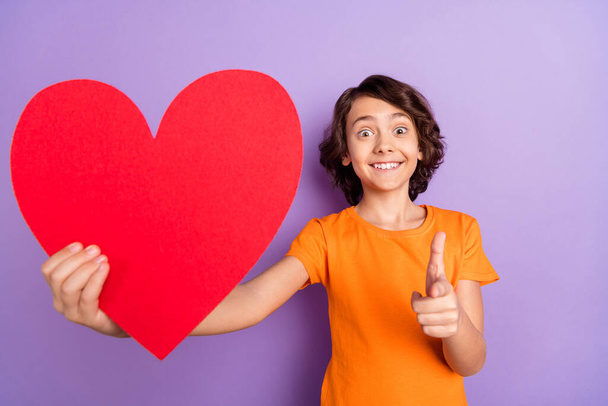 Portrait of nice amazed cheerful boy holding big large heart affection pointing at you isolated over violet purple color background - Foto, imagen