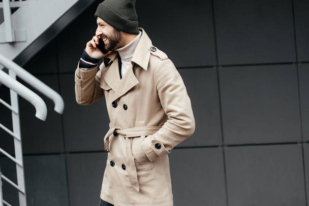 Close-up of a young, smiling, happy, laughing hipster with a beard in a raincoat and hat, talking on a phone and looking at the camera - Photo, Image