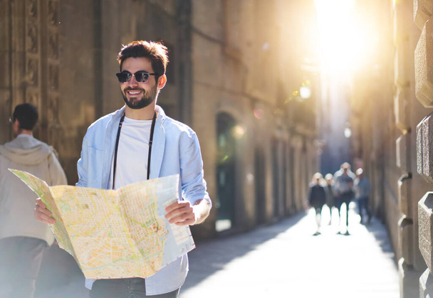 Positive young tourist in casual clothes walking along street with paper map looking for direction in city on sunny summer day - Photo, image