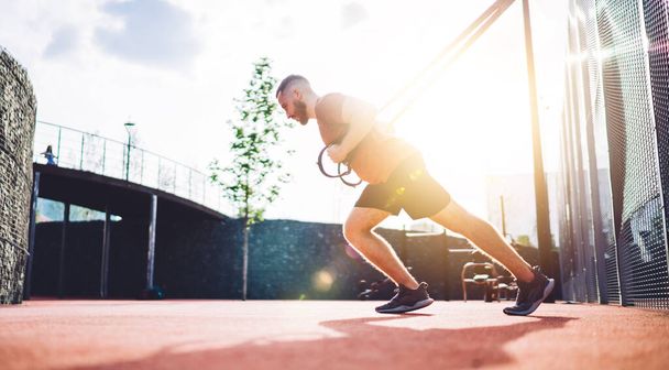 Side view of full body muscular sportsman exercising on TRX straps and looking away while training during morning in sports ground - Fotoğraf, Görsel
