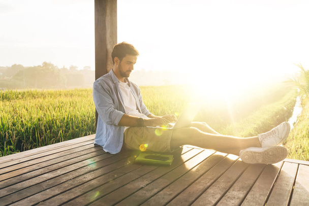 Seitenansicht eines Freiberuflers in Sommerkleidung, der an einem sonnigen Tag mit ausgestreckten Beinen auf dem Holzboden im Feld sitzt und ferngesteuert am Netbook arbeitet - Foto, Bild