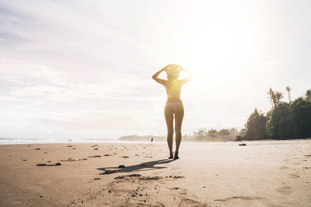 Back view of full body anonymous female in swimwear and hat walking on sandy empty coastline near waving sea during summer holidays - Fotografie, Obrázek