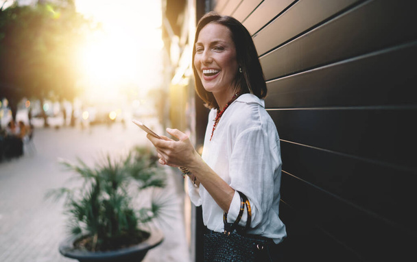 Retrato de mitad de duración de una blogger dentada con tecnología móvil sonriendo a la cámara durante un viaje en solitario, alegre mujer caucásica de 30 años disfrutando del tiempo de llamada durante el ocio para caminar por el día - Foto, Imagen