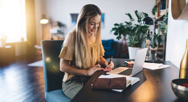 European girl using laptop and writing on adhesive paper at home. Concept of remote or e-learning. Young smiling woman sitting in armchair at desk. Interior of studio apartment - Foto, Bild