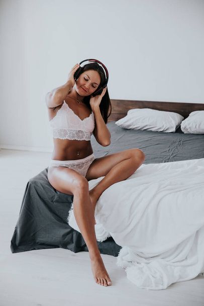 İç çamaşırlı güzel bir kadın yatak odasında kulaklıkla müzik dinliyor. - Fotoğraf, Görsel