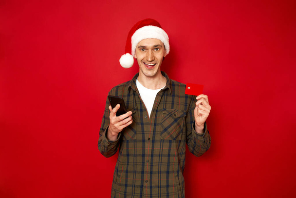 concept of people, online shopping, technology, discounts. an excited man in casual clothes Christmas Santa Claus hat makes an order via phone pays credit bank card, isolated on red studio background - Photo, image