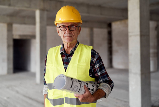 Portrait of caucasian senior engineer holding plans and standing on construction site - Photo, Image