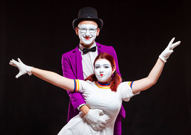 Portrait of two mime artists, isolated on black background. Young woman stands with risen hands, man hugs her from behind, looking at camera - Zdjęcie, obraz