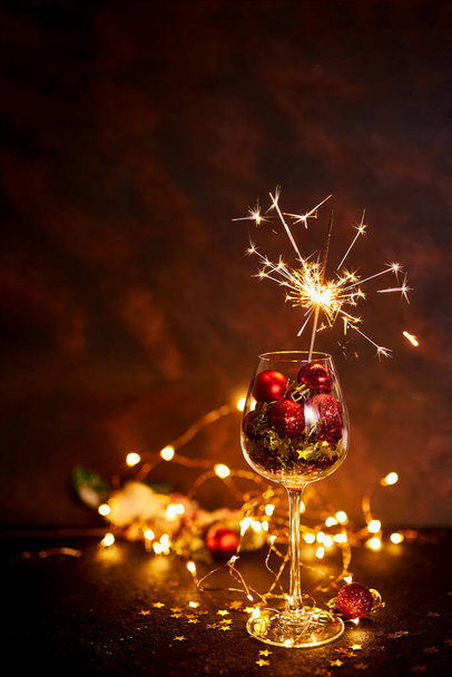 Gelukkig Nieuwjaar. Brandende bengaalse vuurwerk in wijnglas met rode kerstballen op de achtergrond van gouden bokeh lichten. Creatief esthetisch moment. Kerstsfeer en stemming 's nachts. Selectieve focus - Foto, afbeelding