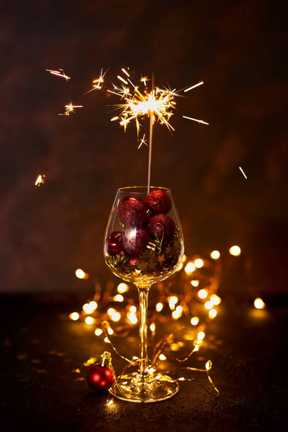Felice anno nuovo. Fuoco d'artificio bruciante bengala in bicchiere di vino con palline di Natale rosse sullo sfondo di luci bokeh dorate. Momento estetico creativo. Atmosfera natalizia e umore di notte. Focus selettivo - Foto, immagini