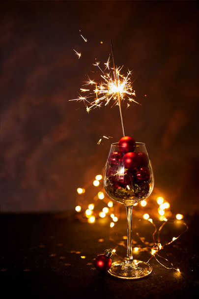 Happy New Year. Burning bengal firework in wine glass with red Christmas balls on background of golden bokeh lights. Creative aesthetic moment. Christmas atmosphere and mood at night. Selective focus - Photo, Image