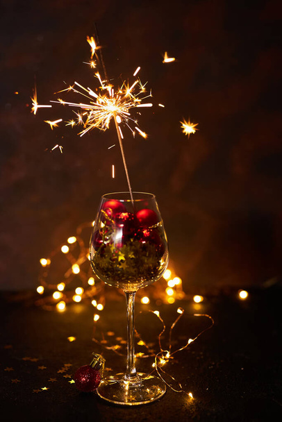 Burning bengal firework in wine glass with red Christmas balls on background of golden bokeh lights. Creative aesthetic moment. New Year atmosphere and mood at night. Selective focus - Photo, Image