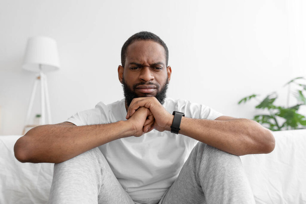Сумний стомлений молодий афроамериканський бородатий чоловік сидить в інтер'єрі кімнати, на білому тлі стіни
 - Фото, зображення