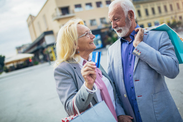 Happy senior couple walking with their shopping purchases on a sunny day using credit card. - 写真・画像