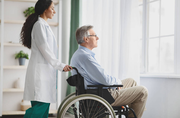 Молодая женщина-врач помогает пожилому мужчине в инвалидной коляске в помещении, копирует пространство - Фото, изображение