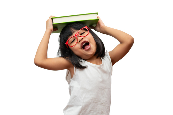 Divertida y feliz asiática niña preescolar con gafas rojas sosteniendo un libro verde en la cabeza, sobre fondo blanco aislado. Concepto de niño en edad escolar y educación en primaria y preescolar, escuela en casa - Foto, imagen
