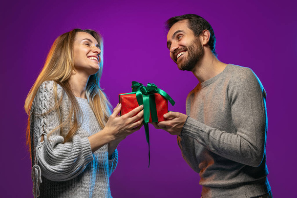 Retrato recortado de feliz pareja joven, hombre dando a la mujer su regalo de vacaciones aislado sobre fondo púrpura en luces de neón - Foto, Imagen