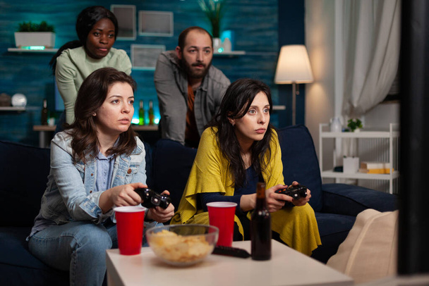 Mujeres concentradas sentadas en un sofá jugando videojuegos usando joystick durante la competencia en línea - Foto, imagen