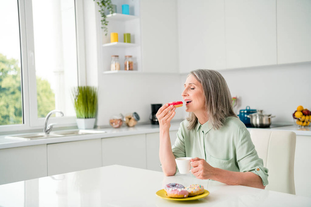 Portrait of attractive elderly dreamy grey-haired woman drinking coffee eating yummy tasty bakery at home light white indoors - Фото, зображення