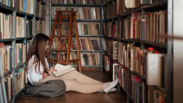 Μια εστιασμένη μαθήτρια κάθεται στο πάτωμα και διαβάζει βιβλίο στη βιβλιοθήκη - Πλάνα, βίντεο