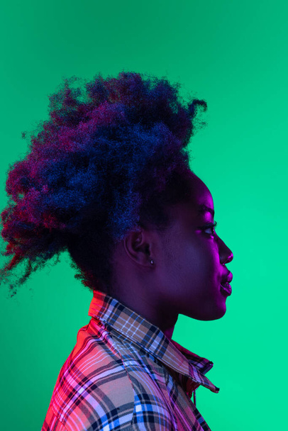 Profile view of dark skinned young girl with adorable hairdo isolated on dark green studio background in purple neon light. - Valokuva, kuva