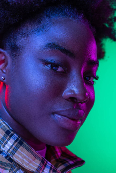 Close-up face of dark skinned young girl looking at camera isolated on dark green studio background in purple neon light. - Фото, зображення