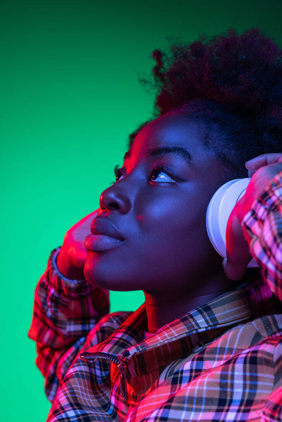 Close-up happy young girl, student listening to music in headphones isolated on dark green studio background in purple neon light. - Foto, Imagen