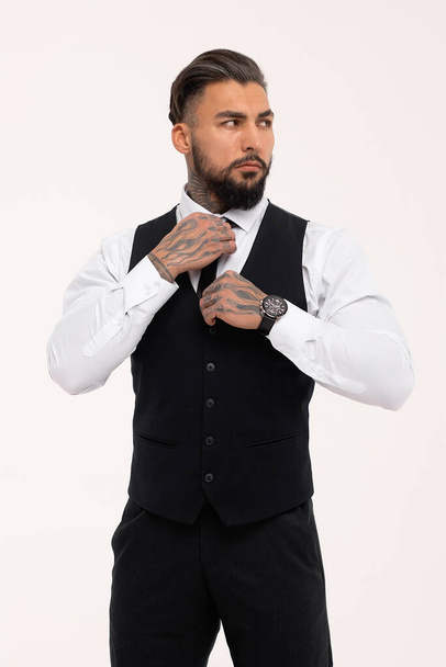 Confident Hispanic bearded man with tattooed hands in white shirt and black trousers adjusting tie and looking away while standing against gray background - Foto, afbeelding