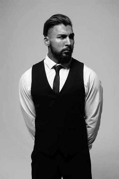 Homem hispânico barbudo em colete escuro e camisa branca com gravata olhando para a câmera com as mãos nos bolsos em estúdio - Foto, Imagem