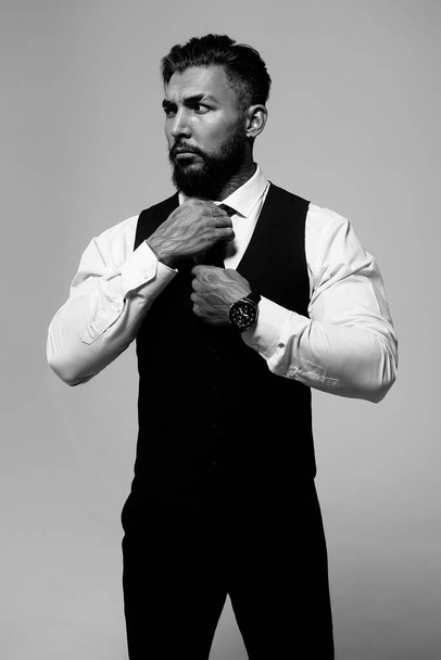 Sebevědomý hispánský vousatý muž s tetovanými rukami v bílé košili a černých kalhotách upravující kravatu a dívá se stranou, zatímco stojí na šedém pozadí - Fotografie, Obrázek