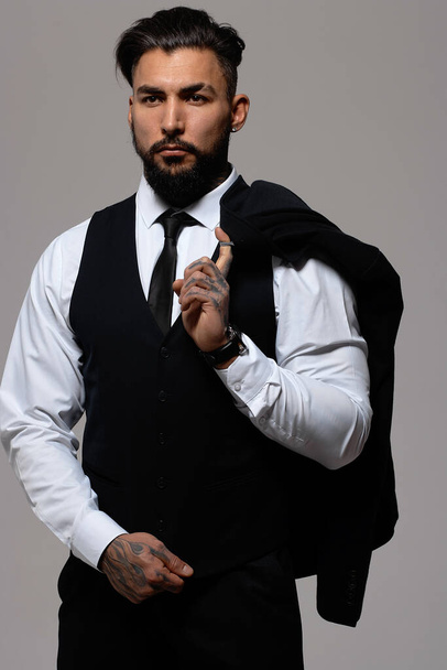 Barbu hispanique gars en gilet sombre et chemise blanche avec cravate regardant caméra avec les mains dans les poches en studio - Photo, image