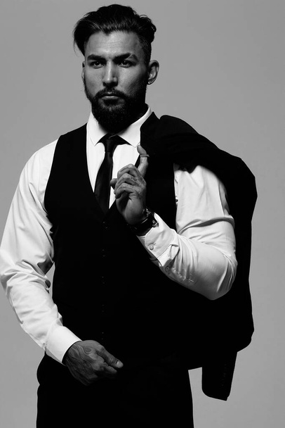 Barbu hispanique gars en gilet sombre et chemise blanche avec cravate regardant caméra avec les mains dans les poches en studio - Photo, image