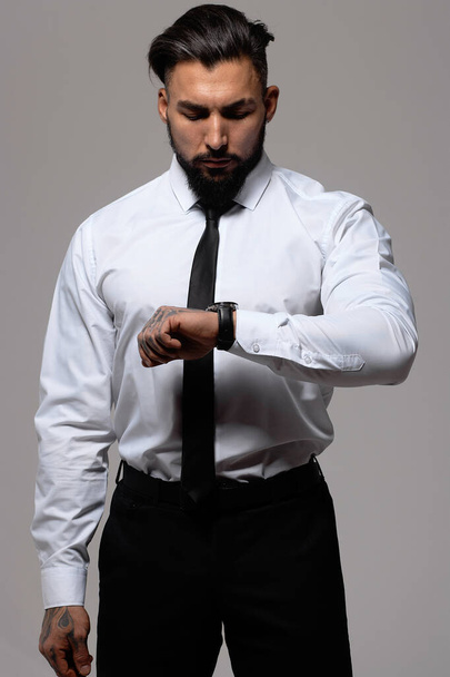 Bearded Hispanic guy in dark vest and white shirt with tie looking at camera with hands in pockets in studio - 写真・画像