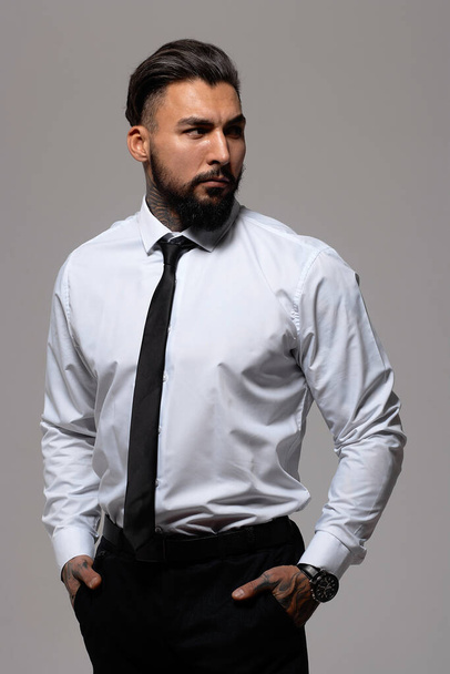 Chico hispano barbudo con chaleco oscuro y camisa blanca con corbata mirando a la cámara con las manos en los bolsillos en el estudio - Foto, imagen