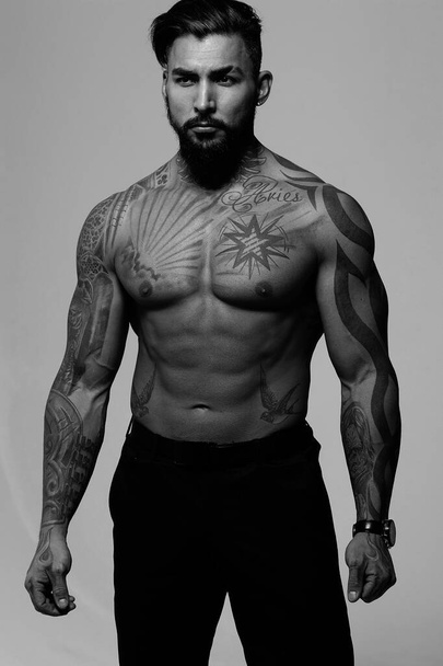 Modelo masculino sem camisa hispânica com tronco tatuado muscular de pé com as mãos nos bolsos e olhando para o fundo cinza - Foto, Imagem