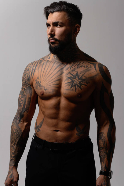 Modello maschile ispanico senza maglietta con busto muscoloso tatuato in piedi con le mani in tasca e distogliendo lo sguardo sullo sfondo grigio - Foto, immagini