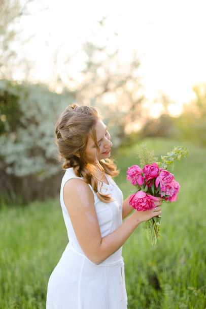 Joven chica caucásica mirando flores de ramo y usando vestido blanco afuera. - Foto, Imagen